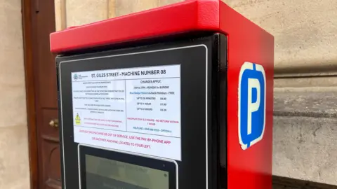 A close up of one of the electronic parking machines, which is painted red on the sides and top. There is a big blue P sign on the side.
On the front there is a white sign showing the charges: £0.80 for up to 30 minutes, £1.60 for up to an hour and £3.20 for up to 2 hours. This one is titled St Giles Street - Machine Number 08