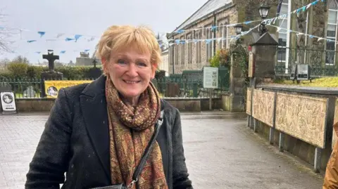 Shows a woman with red hair smiling. She is wearing a multi-coloured scarf, black coat and black handbag. 