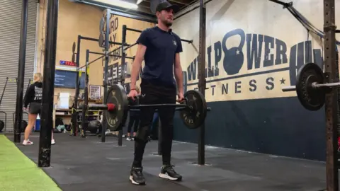 A man with a blue T-shirt and black trousers and a dark cap holding a weights bar in a gym.