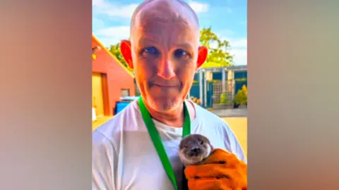 Kevin Murphy A man wearing a white T-shirt and an orange glove holds a young otter on his chest on a sunny day. 