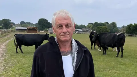 Martin Giles/BBC Ricky Holland with short white hair looking at the camera and wearing a black zip-up jacket over a white top. There are black cattle behind him in a field. There are trees and farm buildings in the background.