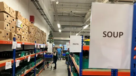 A long aisle in a warehouse stacked with boxes and cans of food. There are signs saying "Pasta" and "Soup". A woman with a trolley is in the distance picking up cans.