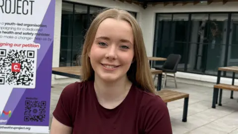 A teenage girl with long blonde hair, sitting at an outdoor bench and smiling at the camera. She is wearing a maroon T-Shirt.