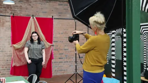 Saturday Town Someone with glasses in a black and white top and black trousers is posing for the camera, which is being held by somebody wearing a yellow top on the right.