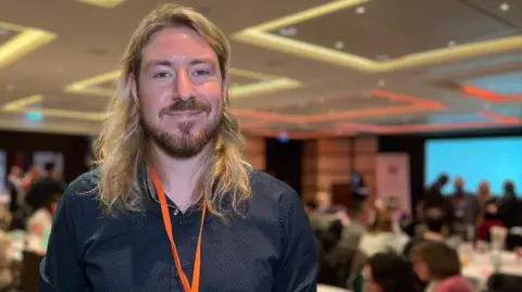 John is wearing a dark shirt and an orange lanyard with a conference badge stands in a large meeting room. In the background, attendees are seated at tables, and presentation screens are visible on the walls.