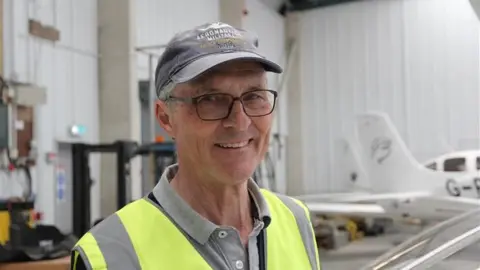 BBC A man in a cap and hi-vis jacket standing inside a warehouse building where aeroplanes are kept.