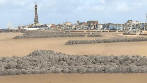 Visualisation of rock headlands along the beach. They are long stretches of grey rocks edging from the steps to the beach towards the sea. Blackpool's coastline can be seen in the distance in the background with the tower to the left
