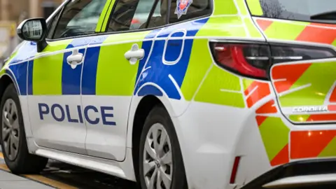 The image shows the side of a police car on the side of the road. Some red barriers can be seen further down the road, blurred in the background.
