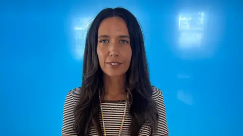 Kathryn looking at the camera. She is wearing a striped brown and white top. She has long brown hair.