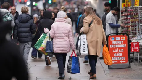 Shoppers wearing coats and carrying bags walk along a damp pavement on Oxford Street in central London.