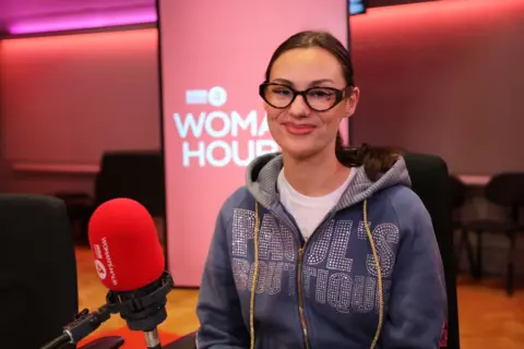 A woman with long hair and glasses wearing a blue hoody sits in the Woman's Hour studio