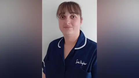 Ollie Dixon A woman in a blue nurse uniform. "Sue Ryder" is written in white on the left side of her uniform.