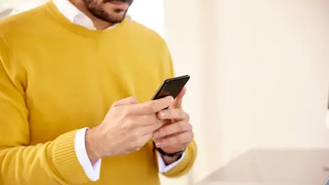 An anonymous man, wearing a yellow jumper, with a white shirt underneath, looking at a phone by a laptop. His face is not visible.