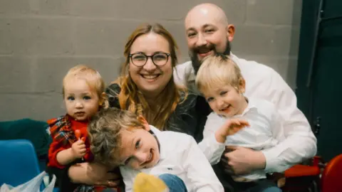 Nikol Biela A mother and father take a photo with three children sitting on their laps - two young boys and a toddler girl. They all smile at the camera.