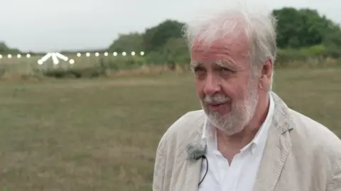 A man with grey hair is wearing a white shirt and beige jackety and is stood at the end of Manchester Airport runway
