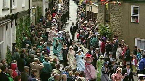 A photo from 1997 shows a procession of couples dressed for the Midday Dance. The women are wearing floor-length gowns with gloves and hats while the men are wearing top hats and tails. There are spectators lining the route and the shops are decorated with greenery.