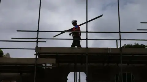 A construction worker in silhouette carries scaffolding poles over his shoulder on a building site
