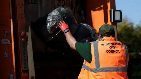 A Birmingham City Council bin worker putting a black bag in the back of a lorry. He has an orange vest on and is wearing a red glove. It is a bright and sunny day.