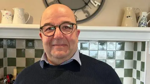 Councillor Mustapha is wearing glasses and a blue sweater and stripped shirt. In the background there are green and white tiles, a large clock and a shelf with porcelain jugs.