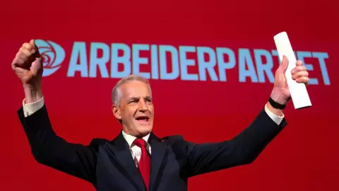 Carl Court/Getty Images A man in a dark jacket and red tie lifts in arms in celebration in front of a red background