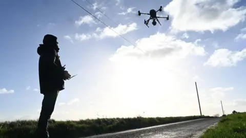 National Grid/StirlingX A drone flying above a road, operated by a person. Both are in silhouette, with a blue sky with clouds. 
