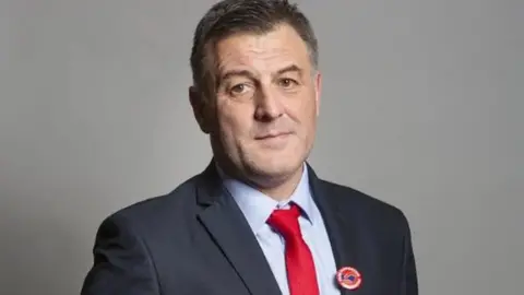 UK Parliament Ian Byrne has short brown hair and is wearing a navy suit with a light blue shirt and a red tie.