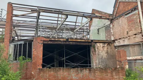 A derelict building with lots of exposed roofing beams and scaffolding that looks like it's supporting the inside of the building. Walls are completely missing in places.