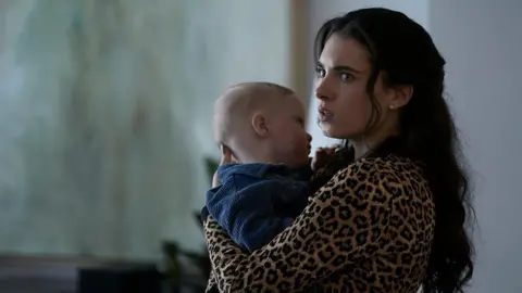 BBC/Left Bank Pictures/James Stack A young woman with long dark hair and wearing a leopard print top is pictured inside a room holding a baby dressed in a blue corduroy shirt.
