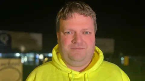 Elizabeth Baines/BBC Parent Matt Ford wears a bright yellow "save our school again" hoodie.