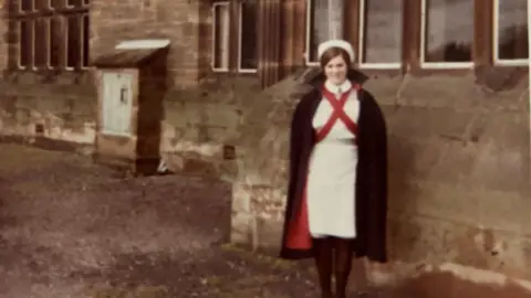 Family Beverley Pedley, in a white dress and black cape with red lining, is stood next to a large stone building which appears to be a hospital.