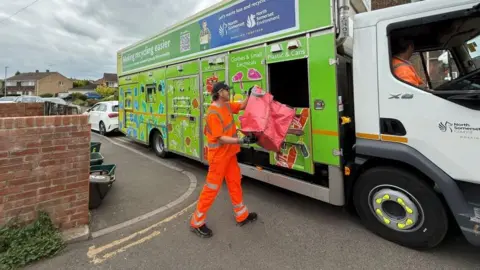 North Somerset Council A man dressed in florescent orange is loading recycling into a green council recycling van. 