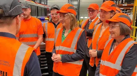 Rachel Reeves wearing orange hi-viz in the centre of the photograph, surrounded by workers a train depot to her left and politicians to her right, including Jo Stevens and Mick Antoniw. All are wearing protective eye wear.