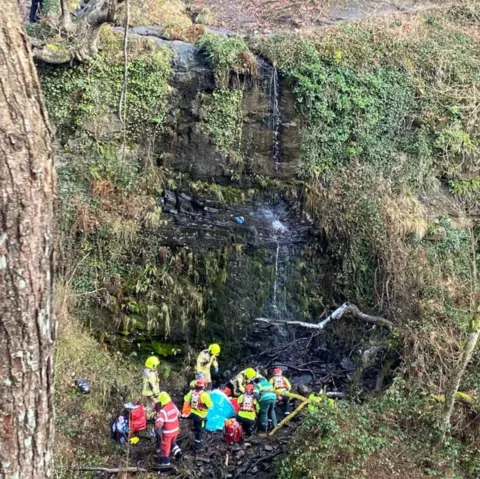Central Beacons Mountain Rescue Team An outdoor scene of rocks and greenery shows a ledge at the top with a small waterfall trickling off.  At the bottom of the photo, a group of mountain rescuers and firefighterss are huddled around someone who has been covered by a blanket.