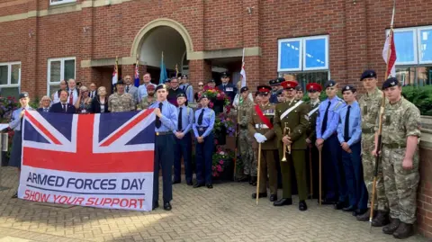 Members of the armed forces gathered at the flag-raising ceremony in Grantham. The personnel are standing behind a union flag with the wording "Armed Forces Day - Show Your Support". They are standing in front of a large red-brick building with arched architectural features and a crest mounted above the entrance.