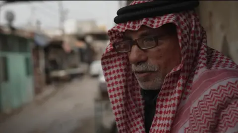 Maher stands in a street wearing a headscarf and glasses 