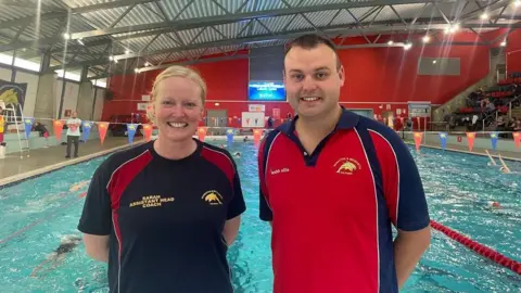 A man and a woman are standing in front of a blue swimming pool with people swimming in the background. The man has short brown hair with a red and navy swimming club t-shirt on. The woman has blonde hair tied in a ponytail and is wearing a black and red swimming club t-shirt 