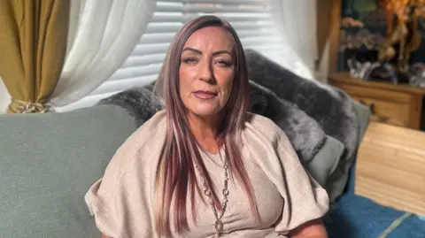 Julieanne Boyle sitting on a green sofa in a living room looking directly at the camera.  She has long, straight hair with blonde and copper tones. She is wearing a loose beige top, and a long silver and pearl necklace. 
