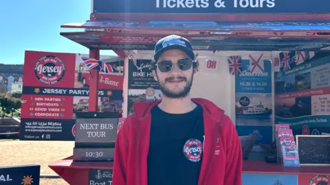 Man in cap and sunglasses and red hoodie in front of a tourism business stand