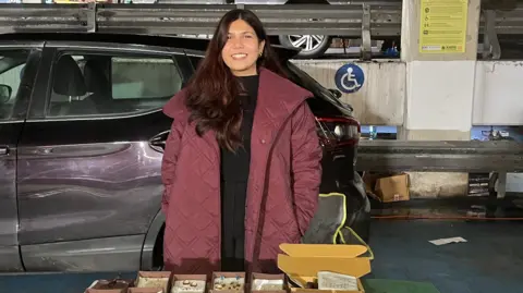 Abeer Laiq has long black hair and is wearing a black top and maroon winter coat. She is standing behind a table next to a car and has jewellery laid out in boxes. She is smiling at the camera.