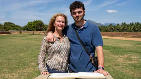 BBC/Studio Lambert Tom and Caroline stood together next to the sign-in book which is the finish line for Race Across The World. They have their arms around each other.