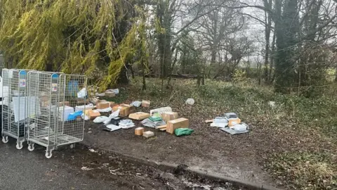 Wes Two empty parcel cages and dozens of packages scattered on a muddy verge surrounded by trees. 