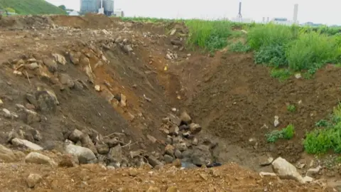 BBC Some rubble in a hole surrounded by some grass on the right and dirt on the left. 