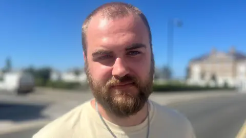 A young man with short auburn hair and a beard looking straight at the camera. He is not smiling and he is wearing a yellow tshirt and a silver necklace. The sky is very blue and there is an out of focus road behind him.