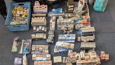 BBC Hundreds of packets of cigarettes and tobacco laid out in piles on an office carpet. 