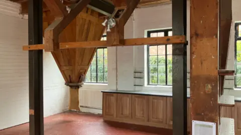 A large suspended wooden cone shaped structure points towards the ground, with a hessian sack covering the end. It is in the corner of a large room with white walls, black framed windows and a red floor.