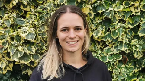 Countryside Regeneration Trust Helen Darragh is standing in front of a leafy bush, the leaves are green and yellow. She has long brown and blonde hair, is smiling at the camera and is wearing a black hoody. 