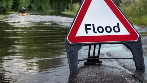 Dorset Council A triangular Flood sign next to a waterlogged area. Someone is seen on a kayak sailing away from it.