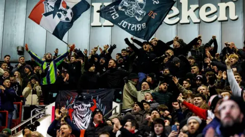 Rogan Thomson/Bristol City FC Bristol City fans, many with their arms aloft, cheer their team on against Stoke City at Ashton Gate. Some are also waving flags with the club's emblem on