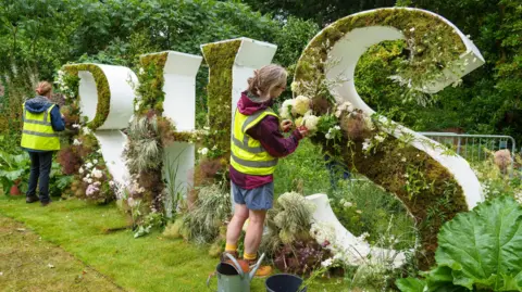 Two people are stood wearing hi-vis jackets in front of large letters which read RHS. They are putting flowers into the letters which are surrounded by different plants.
