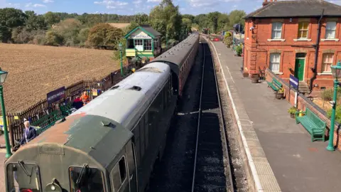 Henry Godfrey-Evans/BBC A bird's eye view of the Mark 1 train stationed in North Weald on a sunny day. There are green wooden benches and lamp-posts.